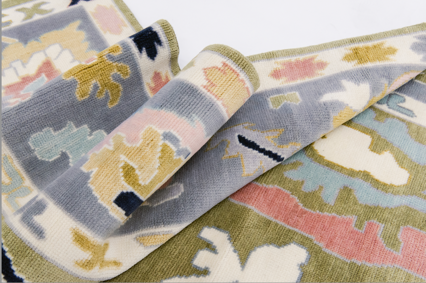 A close-up view of a Penny Modern Oushak rug showcasing its hand-knotted wool texture and the oversized Oushak motifs in various colors including blush, navy, and pale lilac on an olive green background.